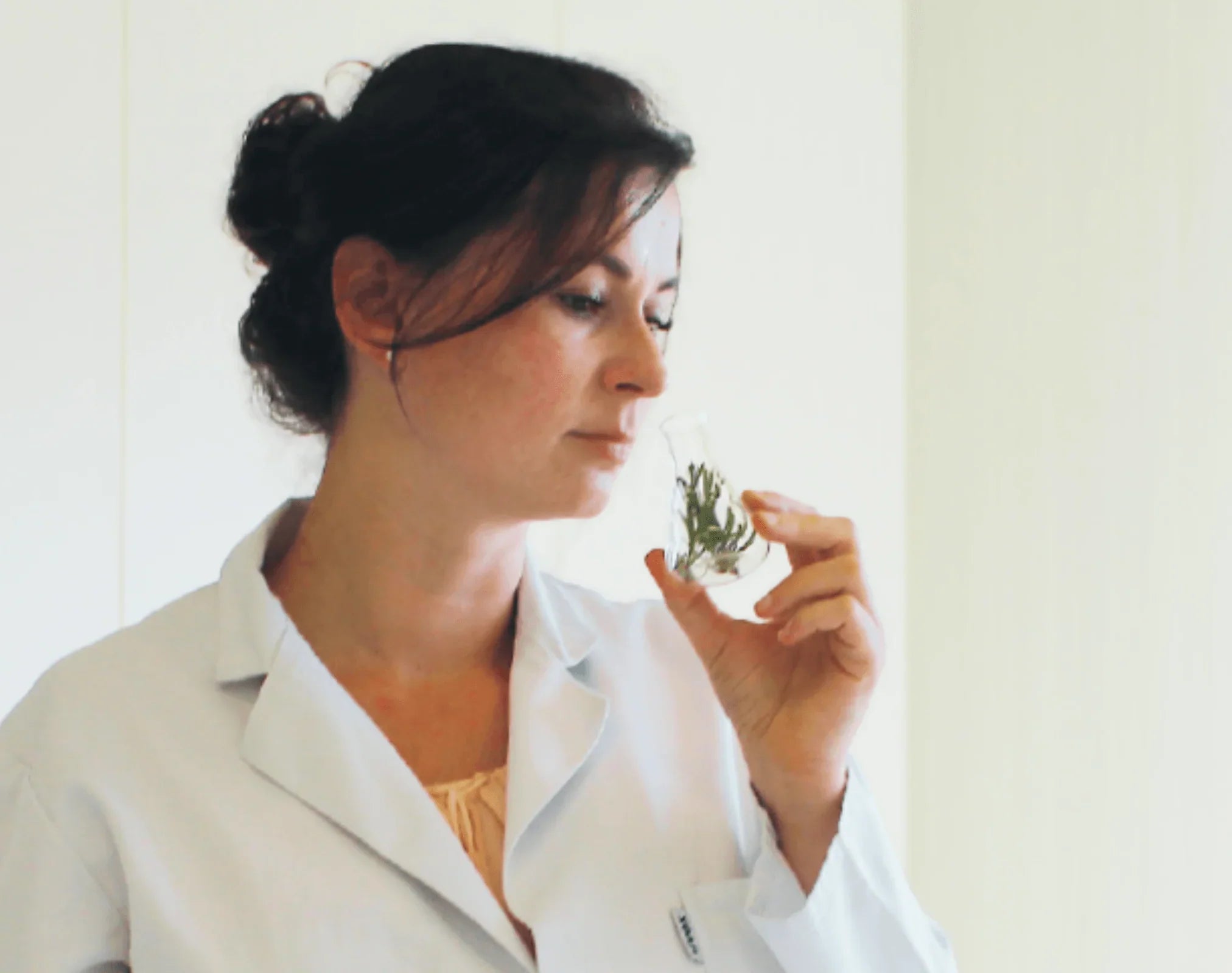 Frau im weissen Laborkittel riecht an einem Glasgefäss mit Pflanzenmaterial; vor ihr stehen Lavendelblüten und Laborgefässe